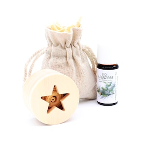Può includere: Un diffusore di legno a forma di stella con un rotolo di legno imbevuto di olio essenziale all'interno. Sono anche raffigurati un piccolo sacchetto di juta marrone pieno di trucioli di legno e una bottiglia di vetro di olio essenziale etichettata "Bio-Alpenzirbe".