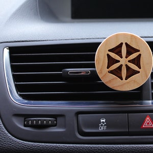 Könnte beinhalten: Ein hölzerner Autolufterfrischer mit geometrischem Design. Der Lufterfrischer ist braun und hat eine runde Form. Er ist an der Lüftung eines Autos befestigt.