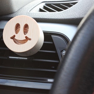 Op de afbeelding: Een houten autoluchtverfrisser in de vorm van een smiley. De luchtverfrisser is lichtbruin en heeft een natuurlijke houtnerf.
