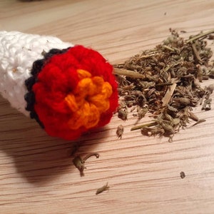 May include: A crocheted catnip toy shaped like a cigarette with a red, orange, and black flower on the end. The toy is filled with dried catnip.