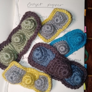 Könnte beinhalten: Fünf gehäkelte Fidget Popper in verschiedenen Farben und Mustern. Die Popper sind aus Garn in den Farben Grau, Blau, Braun, Gelb und Grün gefertigt. Die Popper sind auf einer weißen Oberfläche angeordnet, über ihnen steht der Text "Fidget Popper".