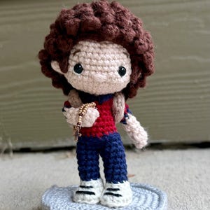 Könnte beinhalten: Handgemachte Häkelpuppe mit braunen Locken, trägt ein rot-blaues Outfit und hält einen Rosenkranz. Die Puppe steht auf einer grauen gehäkelten Basis. Die Puppe hat schwarze Augen und weiße Schuhe.