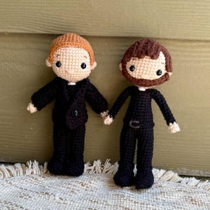 May include: Two handmade crochet dolls holding hands. One doll has light brown hair and is wearing a black suit with a buttoned jacket. The other doll has brown hair and a beard, wearing a black jumpsuit with a belt. Both dolls have black eyes and are on a cream-colored surface.