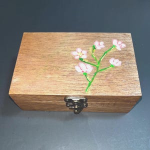 May include: A rectangular wooden box with a hinged lid featuring a hand-painted design of a green stem with four light pink flowers. The box is secured with a silver-toned metal latch on the front, showcasing a natural wood grain finish.