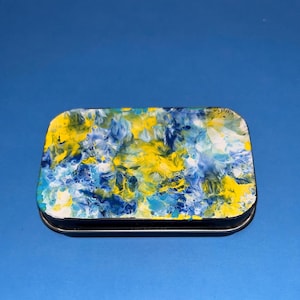 Puede incluir: Una caja metálica rectangular con un diseño abstracto vibrante en tonos amarillos, azules y blancos. La tapa presenta un patrón arremolinado y los bordes son plateados. La caja está sobre un fondo azul liso.