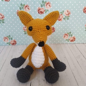 May include: A handmade crochet fox toy with a vibrant orange body, white chest, and black accents on the paws and ears. The fox has large black eyes and a black nose. The toy is sitting on a light wood surface.