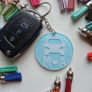 Può includere: Un portachiavi per auto azzurro con una grafica di auto bianca, le lettere "M", un cuore e "A". Il portachiavi è attaccato a una chiave dell'auto e circondato da nappe colorate.