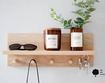 May include: A light wood wall mounted shelf with three hooks and a small shelf. The shelf has a candle in a brown glass jar with a cork lid, a vase with green leaves, sunglasses, and a set of keys hanging from the hooks. The text on the candle jar reads "RETRO Amber & Tonka" and the text on the vase reads "HAPPINESS THERAPY". The text on the bottom right corner of the shelf reads "oh. my wood."