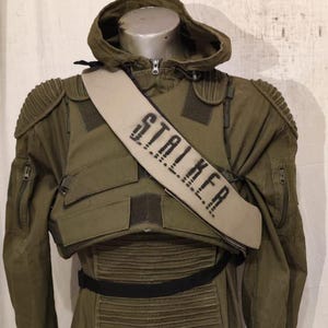 Può includere: Tuta militare verde oliva con cappuccio e imbottitura sulle spalle. Una fascia beige sul petto recita "STALKER" a lettere nere. L'outfit include una cintura nera e tasche multiple.