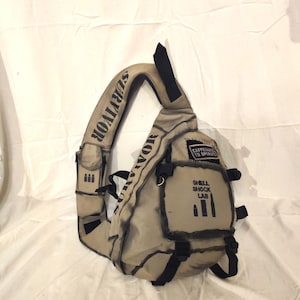 May include: A tan and black shoulder bag with the word "SURVIVOR" printed on the strap. The bag has black accents, straps, and the text "CAFFEINATE TO OPERATE" and "SHELL SHOCK LAB" printed on the front.