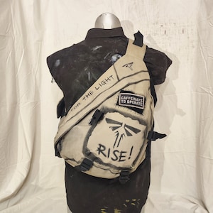 May include: A tan canvas sling bag with black detailing and the phrases "FOR THE LIGHT" and "RISE!". The bag has a patch reading "CAFFEINATE TO OPERATE" and a firefly graphic. It features adjustable black straps and buckles.