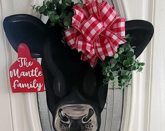 May include: A black cow head door hanger with a red and white gingham bow and greenery. The cow head has a red tag that says "The Mantle Family".