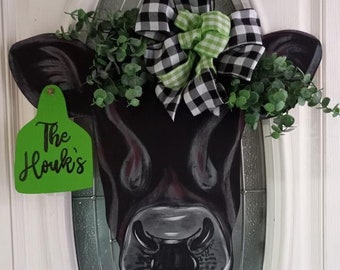 May include: A black and white cow head door hanger with a green wreath and a black and white checkered bow. The sign says "The Houk's".