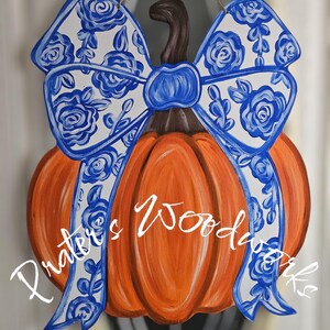May include: Orange pumpkin-shaped wooden door hanger with a brown stem and a large bow. The bow is white with a blue floral pattern. The text "Prater's Woodworks" is written in white script across the pumpkin.