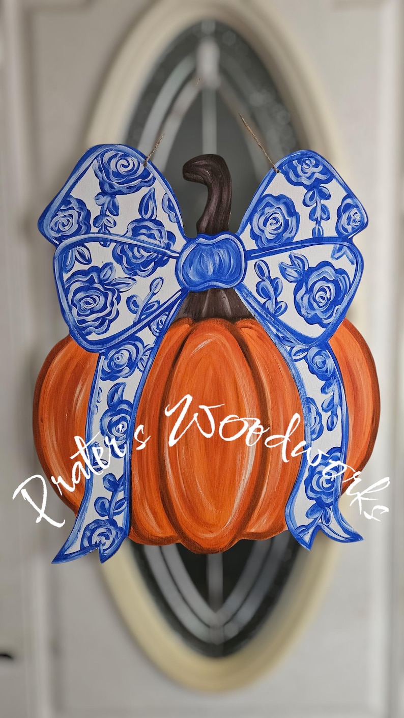 May include: Orange pumpkin-shaped wooden door hanger with a brown stem and a large bow. The bow is white with a blue floral pattern. The text "Prater's Woodworks" is written in white script across the pumpkin.