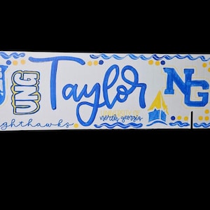 May include: A white rectangular sign with blue and yellow accents. The sign features the name "Taylor" in blue script, along with the letters "UNG" and "NG" in blue. The words "University of North Georgia" are also present.