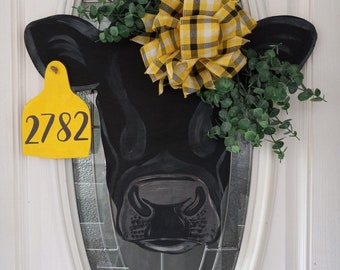 May include: Black cow head door hanger with a yellow plaid bow and greenery. A yellow tag with the number 2782 is attached to the cow's ear.