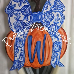 May include: Wooden pumpkin door hanger with a blue and white floral bow and a blue letter "W". The pumpkin is orange with a brown stem. The bow has a floral design. The text "Praters Woodworks" is on the pumpkin.