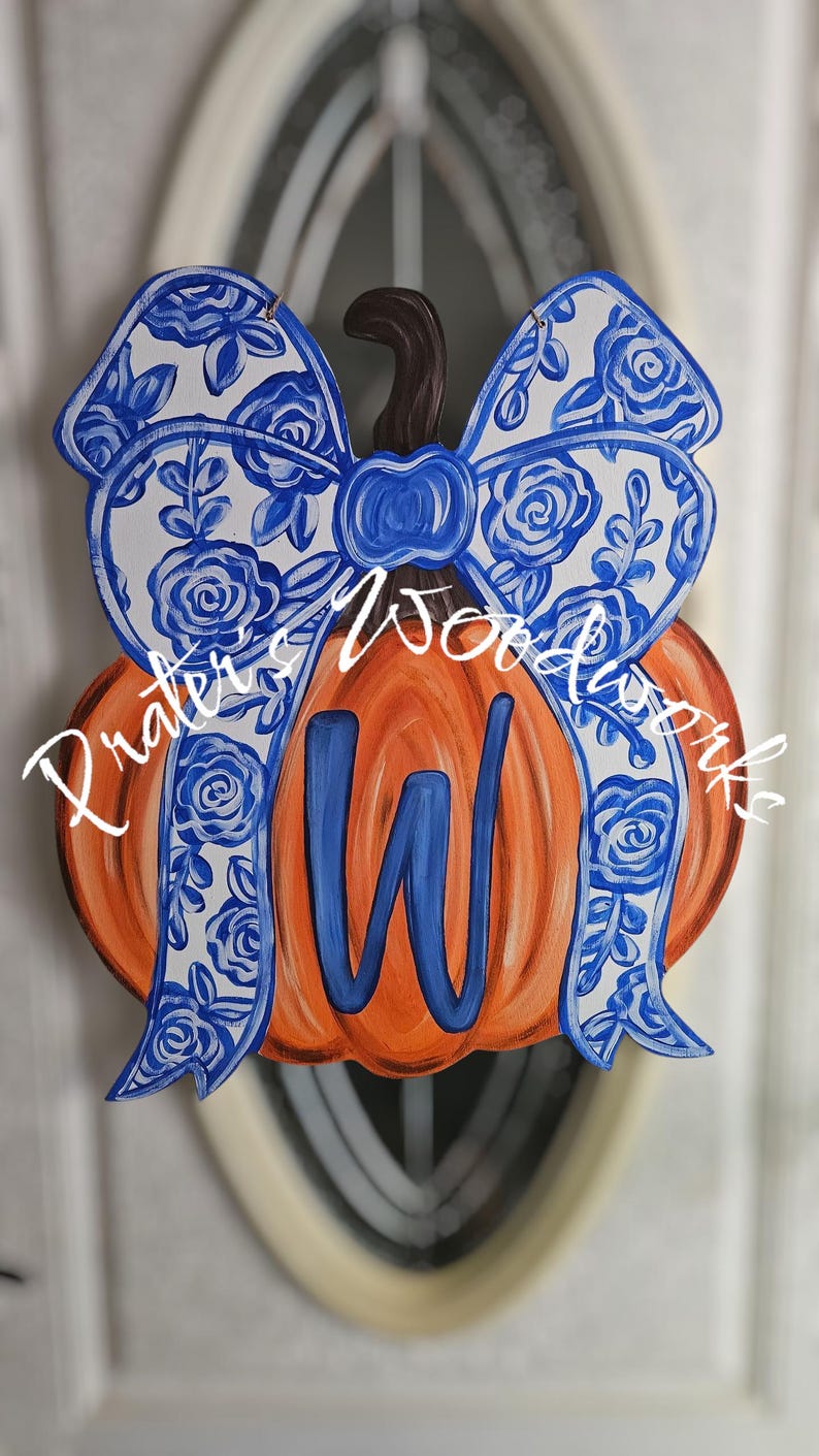 May include: Wooden pumpkin door hanger with a blue and white floral bow and a blue letter "W". The pumpkin is orange with a brown stem. The bow has a floral design. The text "Praters Woodworks" is on the pumpkin.