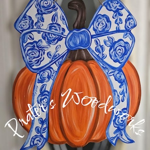 May include: Orange pumpkin-shaped wooden door hanger with a brown stem and a large bow. The bow is white with a blue floral pattern. The text "Prater's Woodworks" is written in white script across the pumpkin.
