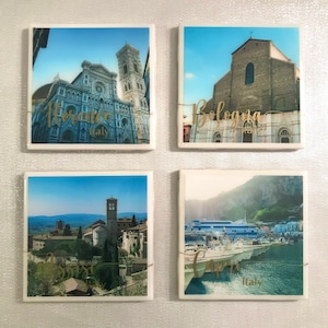 May include: Four square ceramic coasters with images of Italian cities. The coasters feature images of Florence, Bologna, Assisi, and Capri. Each coaster has the city name and "Italy" written in gold lettering.