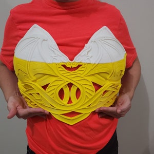 Puede incluir: Un peto decorativo bicolor en blanco y amarillo. La parte superior es blanca con un dise&ntilde;o texturizado en forma de ala. La secci&oacute;n inferior es amarilla con un intrincado patr&oacute;n calado. La pieza la sostiene una persona.