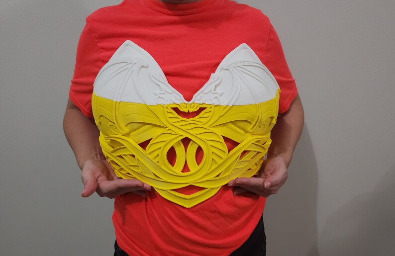 Puede incluir: Un peto decorativo bicolor en blanco y amarillo. La parte superior es blanca con un dise&ntilde;o texturizado en forma de ala. La secci&oacute;n inferior es amarilla con un intrincado patr&oacute;n calado. La pieza la sostiene una persona.