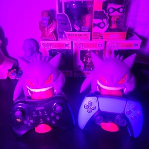 May include: Two purple Gengar character controller stands holding a black Wii U Pro controller and a light-colored PlayStation 5 controller. Behind them are several Funko Pop boxes, including Star Wars Knight of Ren and DC Robin figures.