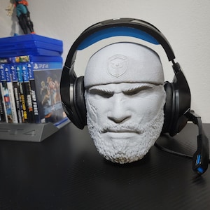 Puede incluir: Un busto gris de un hombre barbudo con gorra blanca con un logo, sosteniendo auriculares para juegos negros y azules. El busto está sobre una superficie negra, con juegos de PS4 y una figura en el fondo.