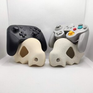 May include: Two video game controllers, one black and one gray, rest on top of matching off-white and speckled gray Cubone skull-shaped controller holders. The controllers are designed for gaming consoles.