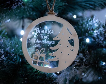 May include: A white wooden Christmas ornament with a clear center. The ornament has a cutout of a Christmas tree and a gift box. The text "Bonnie's First Christmas 2023" is written on the ornament.