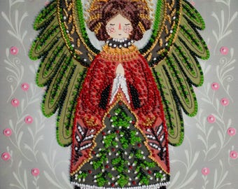 May include: A beaded angel figure with a red robe, green wings, and a gold halo. The angel's dress features a Christmas tree design. The background is grey with a floral pattern and pink accents.