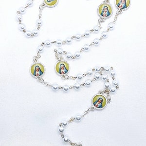 May include: White pearl rosary with silver toned medallions featuring images of the Virgin Mary. The medallions have a yellow background and the Virgin Mary is depicted in blue and white robes.