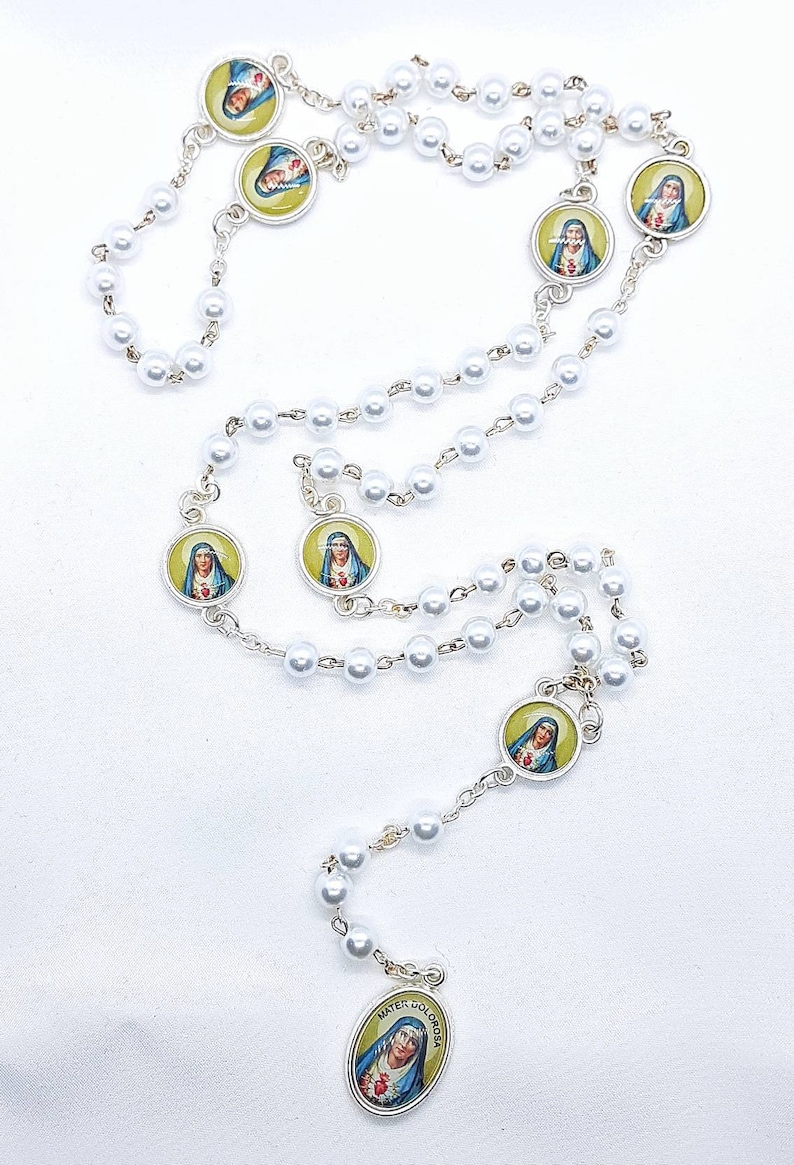 May include: White pearl rosary with silver toned medallions featuring images of the Virgin Mary. The medallions have a yellow background and the Virgin Mary is depicted in blue and white robes.