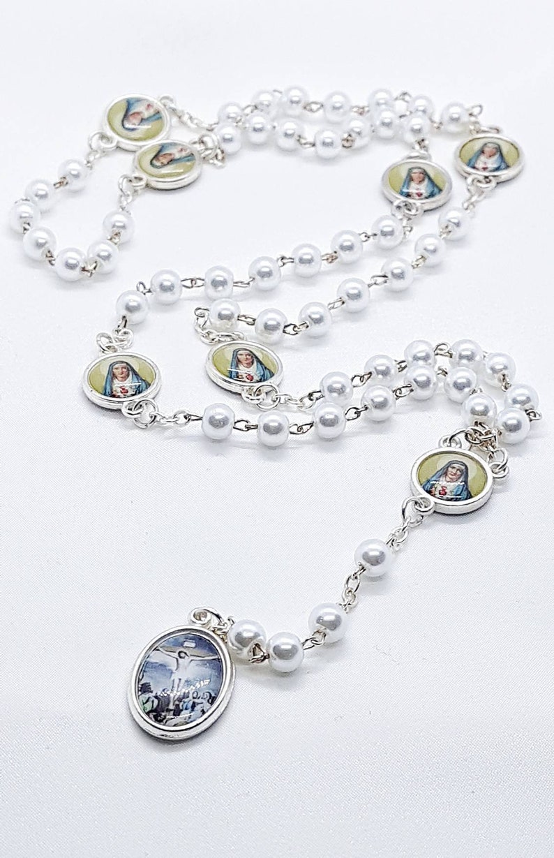 May include: White pearl rosary with silver medallions featuring images of the Virgin Mary and Jesus Christ. The rosary has a crucifix pendant.