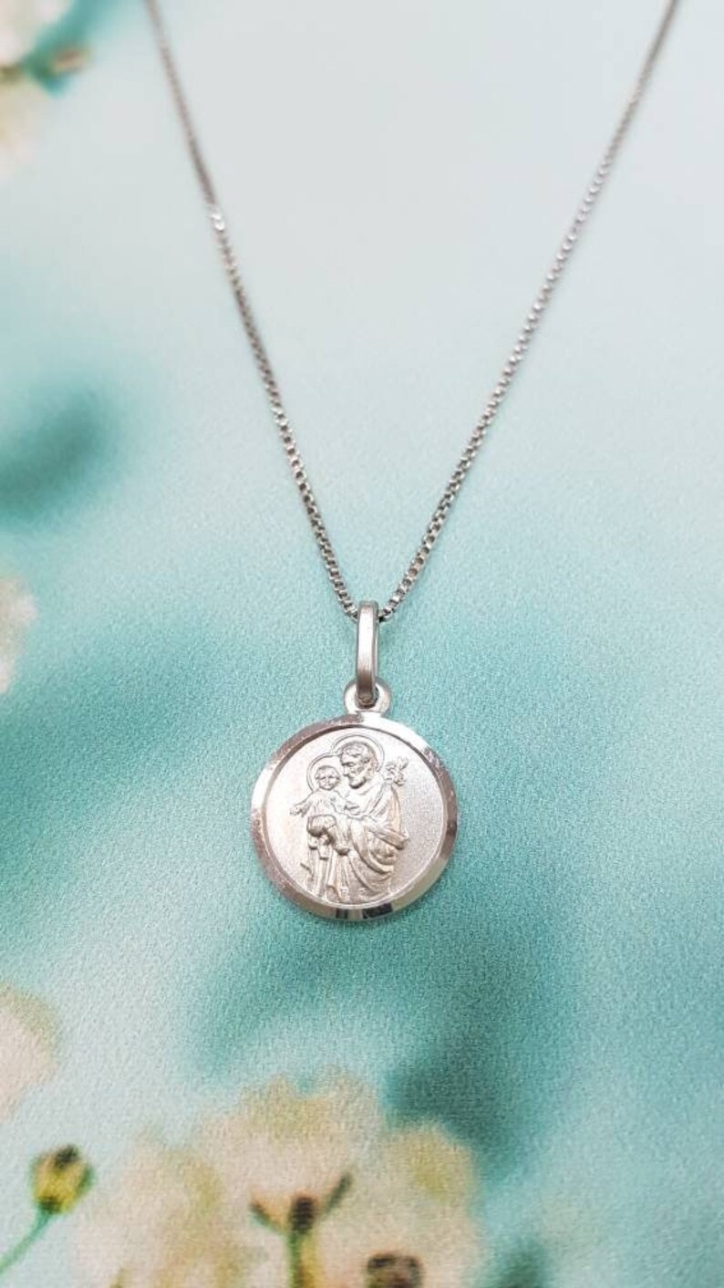 May include: A silver chain necklace with a round pendant featuring a depiction of Saint Joseph holding the baby Jesus.