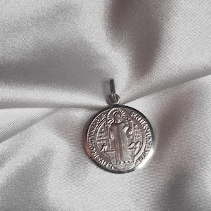 Puede incluir: Un colgante plateado con un diseño circular. El colgante presenta una imagen en relieve de una figura con una túnica, rodeada de texto. El colgante cuelga de un pequeño lazo en la parte superior.