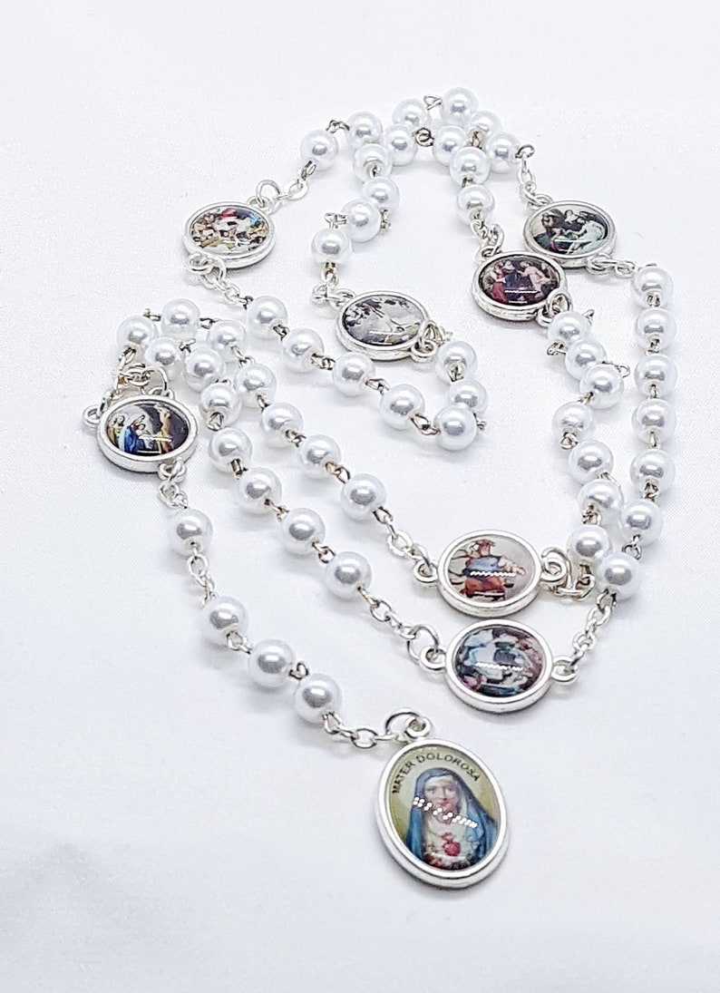 May include: White pearl rosary with silver accents and small round medallions featuring religious imagery. The centerpiece is a silver oval medallion with a depiction of the Virgin Mary.