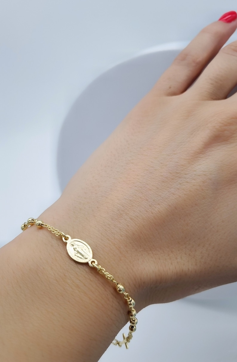 May include: A gold-toned rosary bracelet with a small oval charm featuring a religious image. The bracelet is worn on a person's wrist.