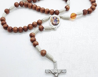 May include: A brown wooden rosary with a white cord and a silver crucifix. The centerpiece features a round image of a religious figure.