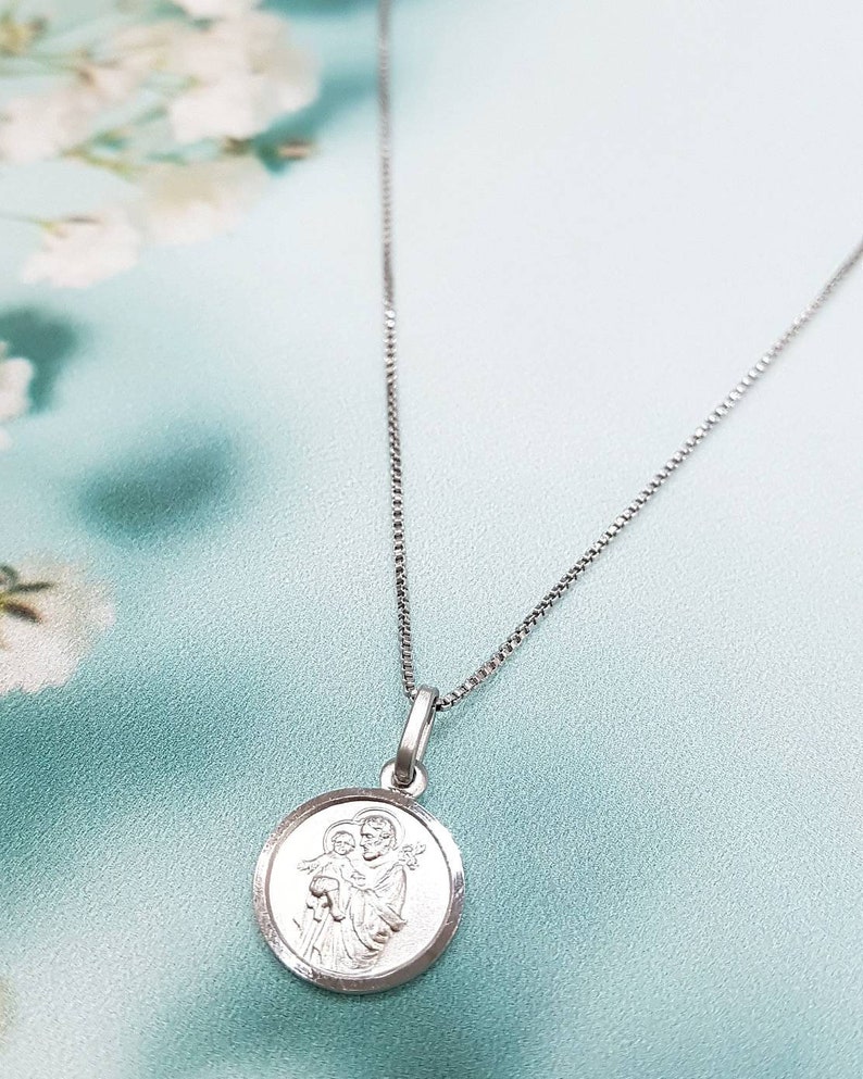 May include: A silver chain necklace with a round pendant featuring a silver image of Saint Anthony of Padua holding a baby Jesus.