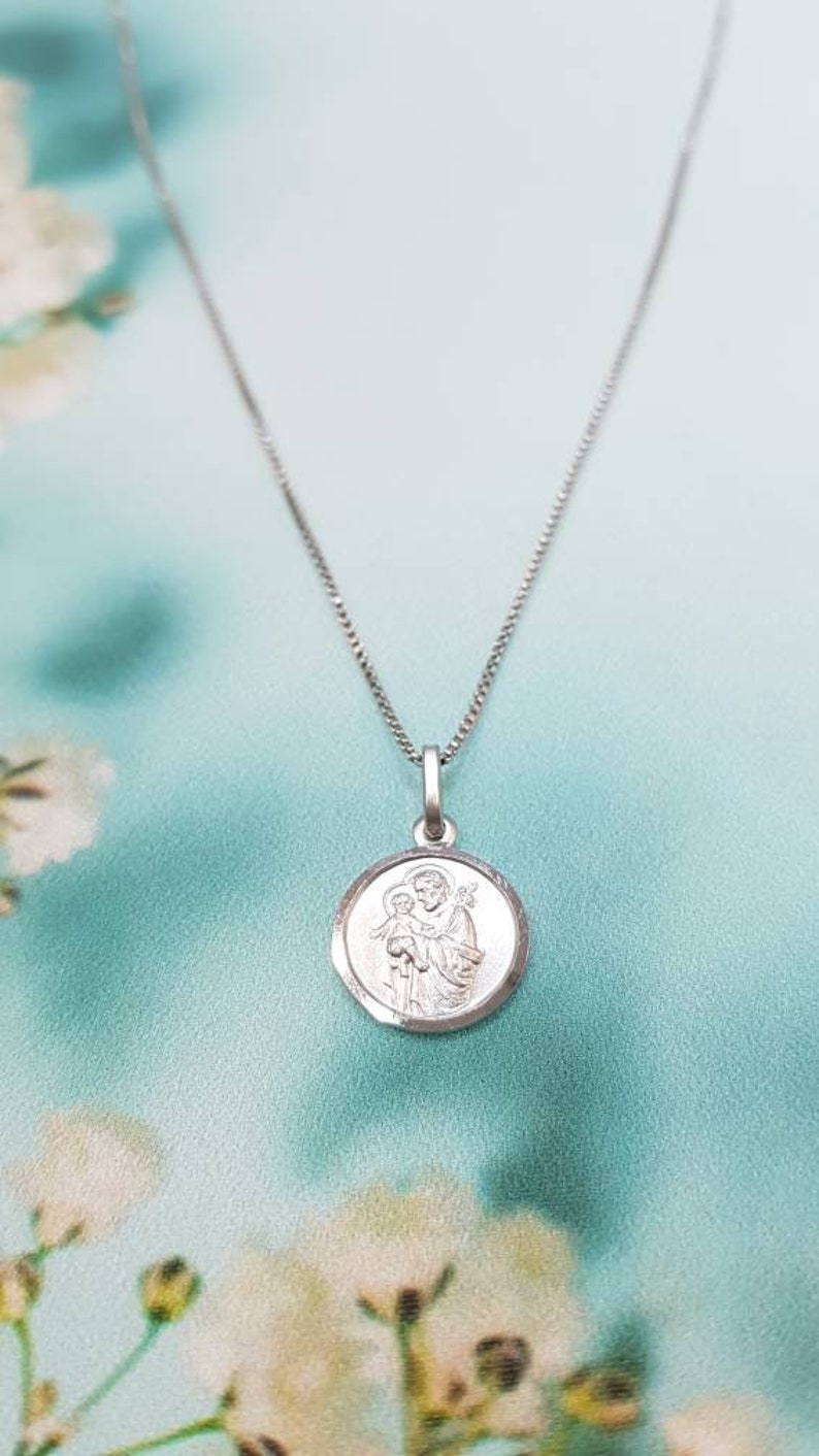 May include: Silver chain necklace with a round pendant featuring a depiction of Saint Anthony of Padua holding a baby Jesus.