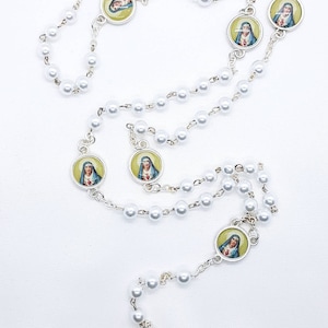 May include: White pearl rosary with silver accents and round medallions featuring images of the Virgin Mary. The centerpiece is an oval medallion with an image of Jesus on the cross.
