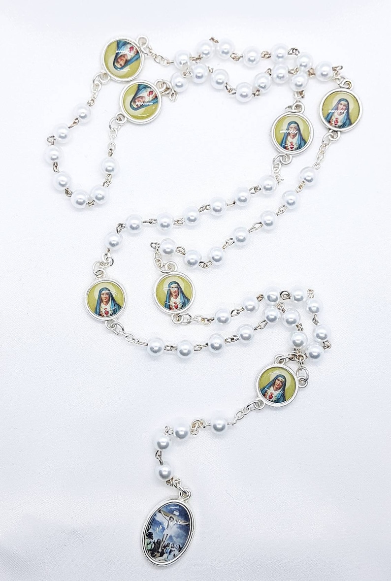 May include: White pearl rosary with silver accents and round medallions featuring images of the Virgin Mary. The centerpiece is an oval medallion with an image of Jesus on the cross.