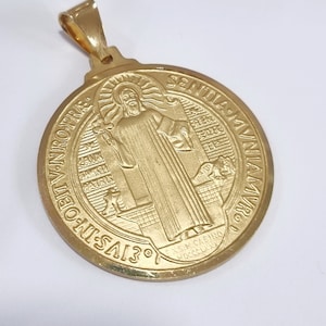 Könnte beinhalten: Ein goldfarbener Anhänger mit einem detaillierten Relief einer religiösen Figur. Der Anhänger hat ein kreisförmiges Design mit Text am Rand und eine Schlaufe für eine Kette. Der Anhänger ist wahrscheinlich aus einem Edelmetall gefertigt.
