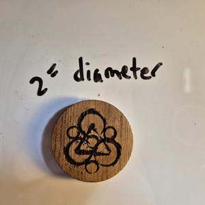 May include: A wooden, circular object with a black, geometric design. The object is 2 inches in diameter. The design features a series of interconnected shapes, including triangles and circles, etched into the wood.