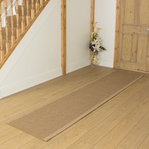 May include: A beige runner rug with a woven pattern is placed on a wooden floor in a hallway. The rug is in front of a wooden door and a staircase with a wooden railing.
