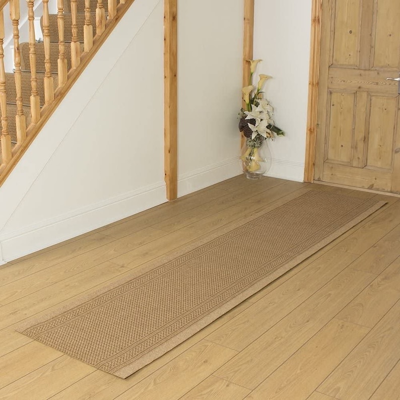 May include: A beige runner rug with a woven pattern is placed on a wooden floor in a hallway. The rug is in front of a wooden door and a staircase with a wooden railing.