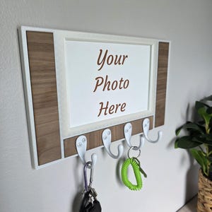 May include: A wooden key holder with a white frame and four white hooks. The key holder has a white center with the text "Your Photo Here" in brown. Keys are hanging from the hooks.