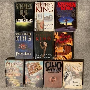 May include: A collection of Stephen King novels, including "The Tommyknockers," "Hearts in Atlantis," and "Nightmares & Dreamscapes." The books feature various cover designs with titles and author's name prominently displayed. The books are arranged on a neutral surface.